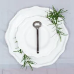Handcrafted stainless steel bottle opener with oxidised dark handle on white ceramic plate, made in Pilimathalawa Kandy Sri Lanka